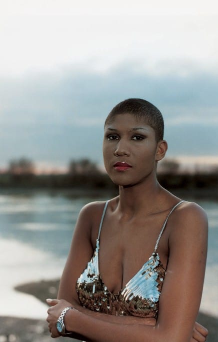 A color photo of a black woman with a close-cropped hair cut, wearing a silver sequined strappy top. She is in front of a body of water.